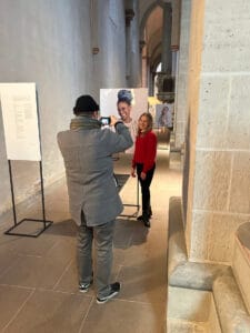 Elena Kaufmann wird neben einer Fotowand, die eine lächelnde dunkelhäutige Frau in weißem Gewand zeigt, fotografiert.