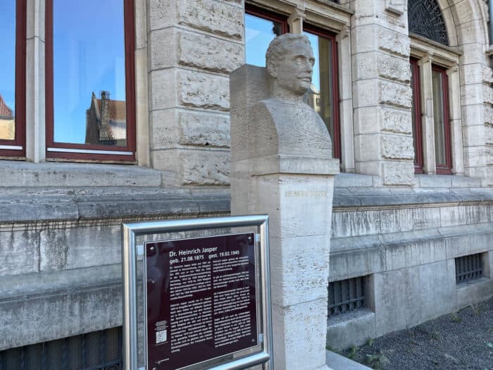 Steinerne Büste von Heinrich Jasper mit Hinweistafel vor einer Steinfassade mit Fenstern.